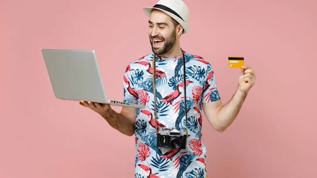 A man in a tropical shirt holds a credit card and laptop, symbolizing travel loans and financial planning for vacations.