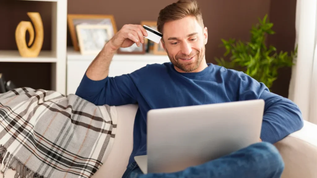A man sitting on a couch using a laptop and holding a credit card for an online loan application.