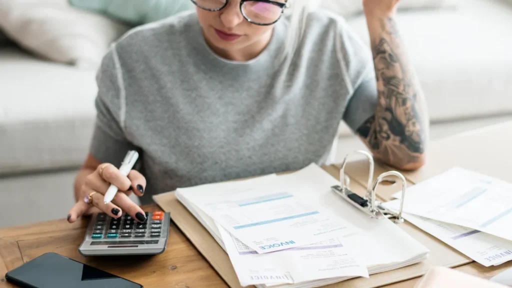 Une femme avec des lunettes utilise une calculatrice à une table, entourée de papiers et d'un téléphone pour un simulateur de prêt.