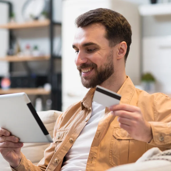 A man sits on a couch, holding a tablet and credit card, exploring options for the best personal loan.