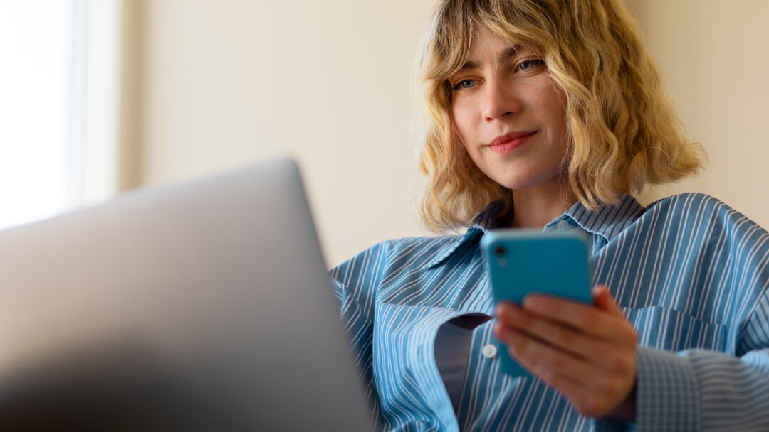 Une femme assise devant un ordinateur portable, tenant un téléphone portable, avec le texte "Prêts numériques au Canada en 2026".